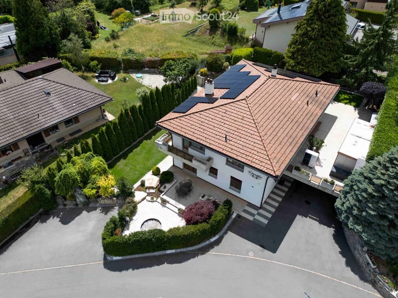 Aerial view of a two-story house with a tiled roof, surrounded by a well-maintained garden, patio, and outdoor seating area. The house features solar panels on the roof and a driveway leading to a garage.