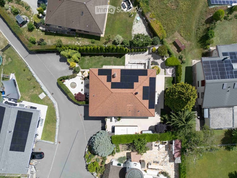 Aerial view of a residential area with multiple houses, solar panels on roofs, gardens, driveways, and parked cars.