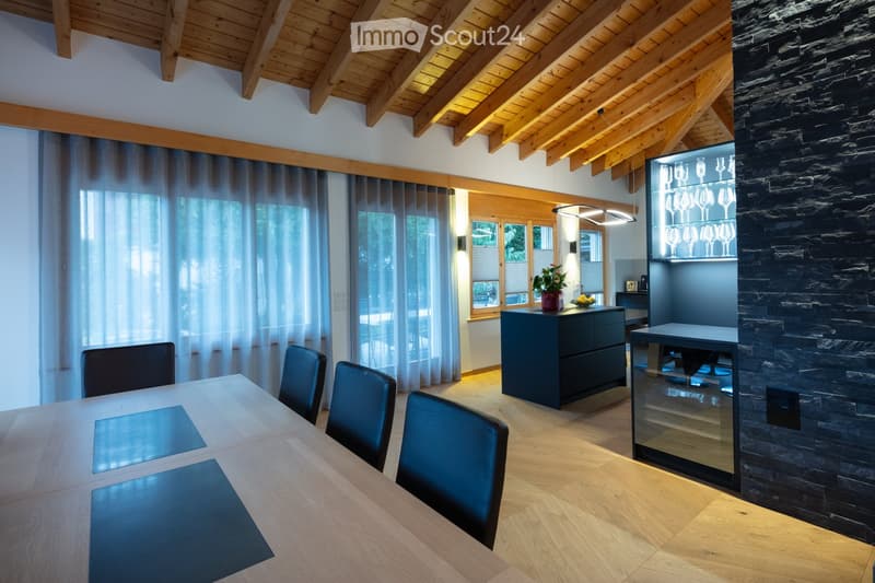 Modern dining area with a wooden table, blue chairs, a dark cabinet, and a wine rack. The room features exposed wooden beams on the ceiling and large windows with sheer curtains.