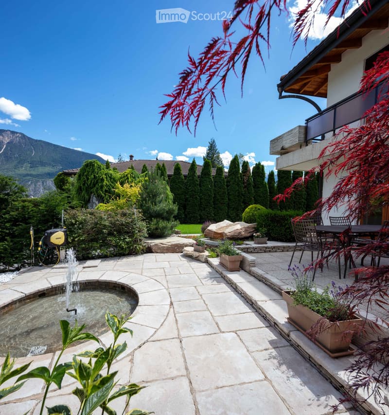 Well-maintained garden with a stone patio, fountain, potted plants, and outdoor furniture, overlooking a mountain view.