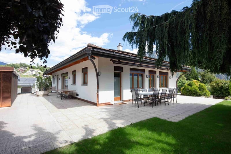 A modern single-story house with a spacious outdoor patio area featuring a dining table and chairs, surrounded by greenery and a well-maintained lawn.