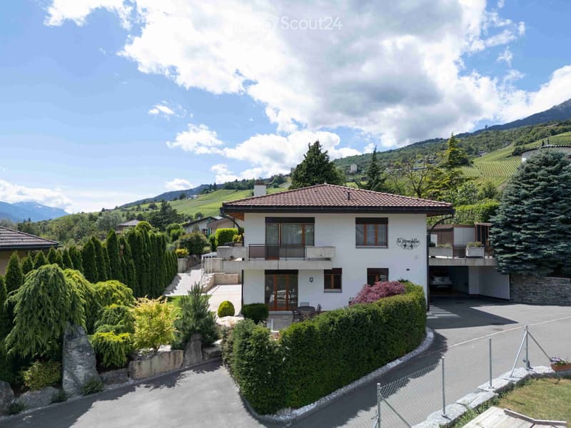 A two-story house with a tiled roof, white exterior, and a balcony on the upper level. The house is surrounded by well-maintained gardens, trees, and a driveway leading to a garage. The background features a scenic view of rolling hills and mountains.