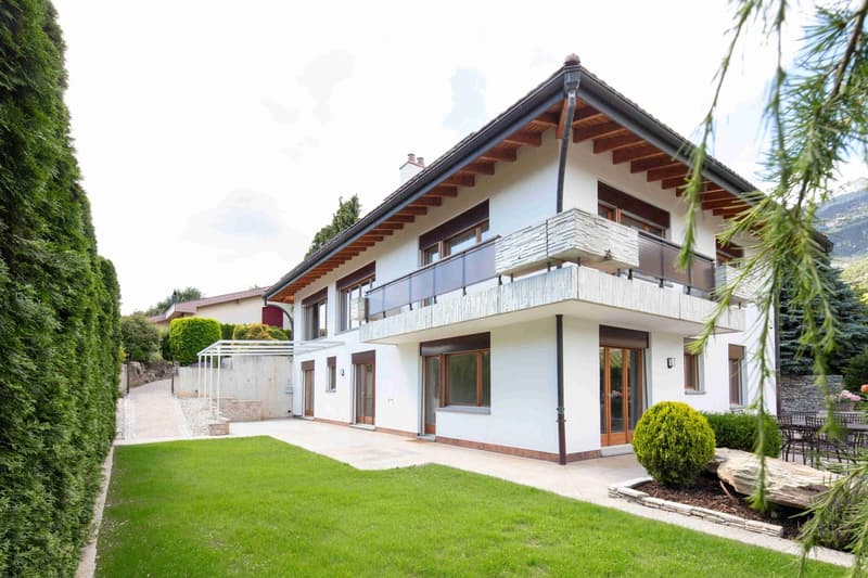 Modern two-story house with white exterior, large windows, and a balcony on the upper floor. The house features a well-maintained lawn, a stone pathway, and surrounding greenery.