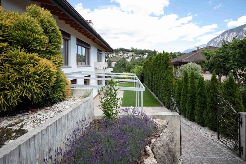 Garden area with a white house, neatly trimmed hedges, lavender plants, a paved pathway, and a view of mountains in the background.