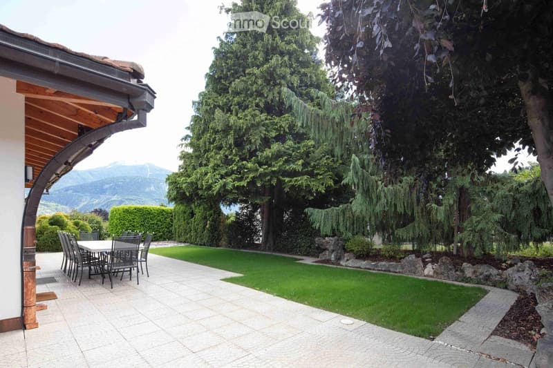 Outdoor patio area with a table and chairs, surrounded by a well-maintained lawn, trees, and a scenic mountain view in the background.