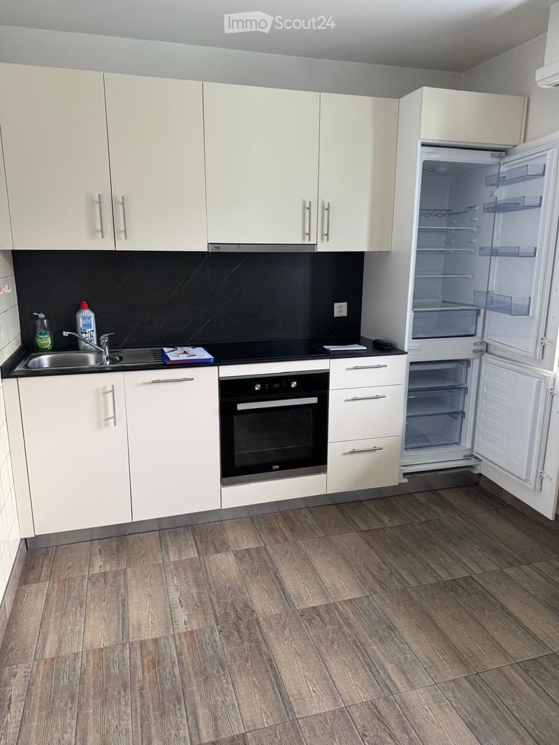 Modern kitchen with light-colored cabinets, stainless steel sink, oven, and open refrigerator. Wooden flooring and black backsplash.