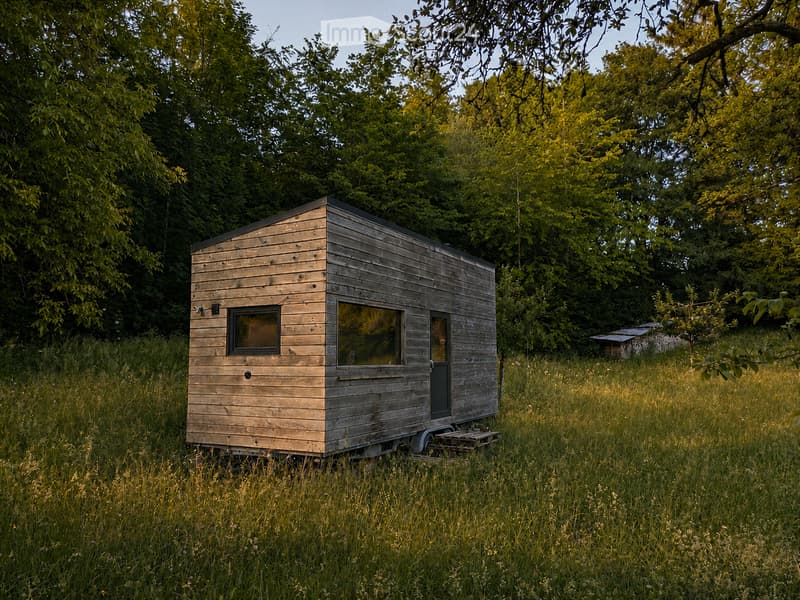 A small, modern wooden cabin with a flat roof, situated in a grassy field surrounded by trees. The cabin features large windows and a door, with a small wooden bench in front.
