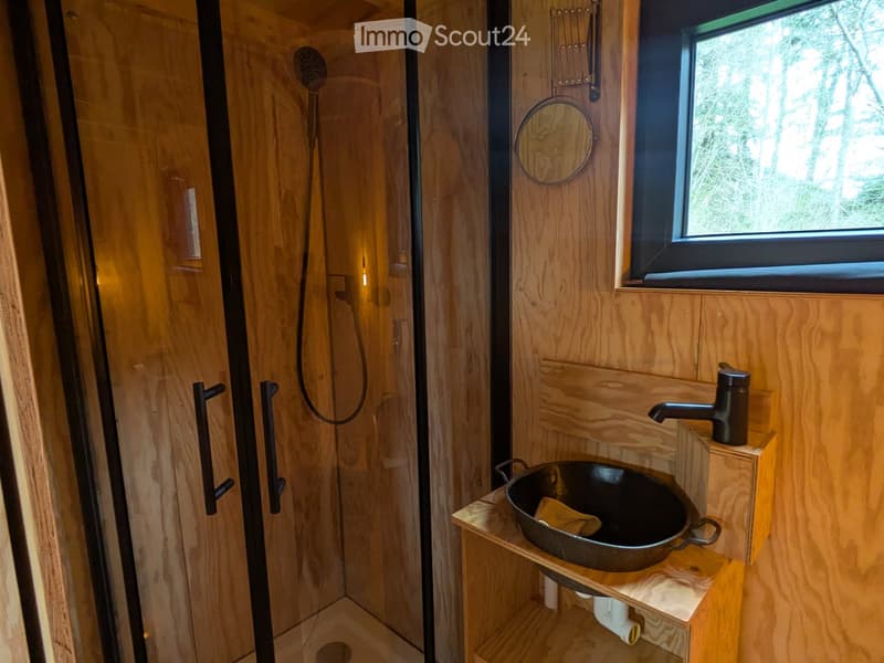 Cozy bathroom with wooden walls, a black sink, a shower area, and a round mirror above the sink.
