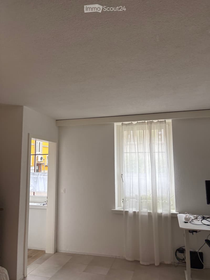 Empty room with light-colored walls, tiled floor, and a window with white curtains. A small table with electronic devices is visible in the corner.