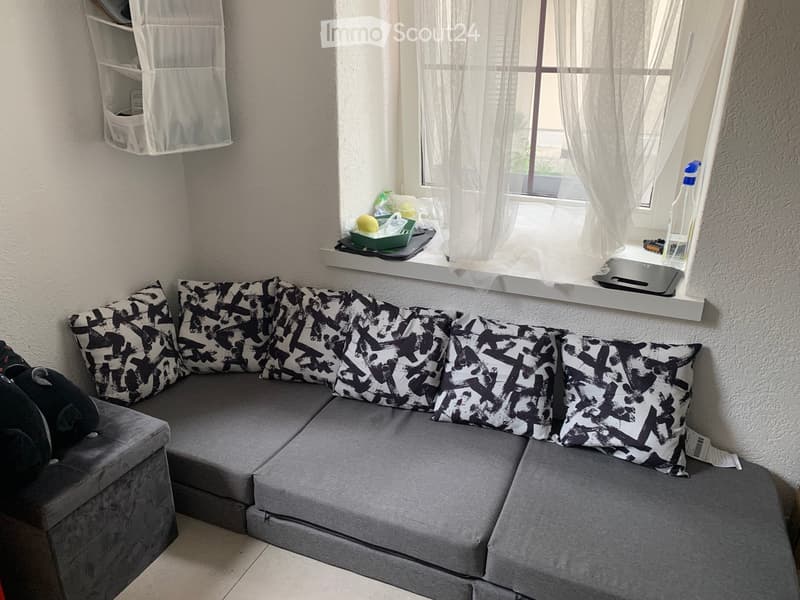 A cozy living room corner with a grey sectional sofa adorned with black and white patterned pillows, a small black stool, and a window with sheer curtains. A small shelf above the sofa holds various items, including a green tray with a yellow fruit and a