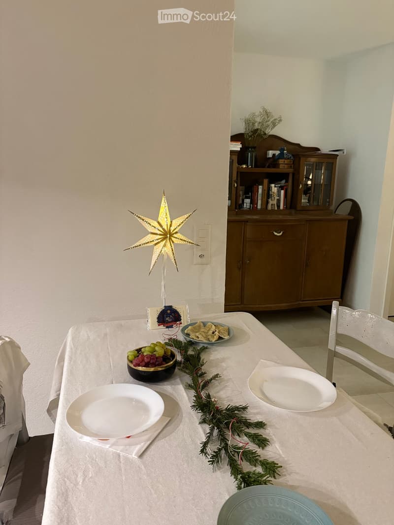 Dining area with a table set for a festive meal, featuring plates, bowls, grapes, and a garland. A wooden cabinet and bookshelf are visible in the background, along with a decorative star ornament on the wall.