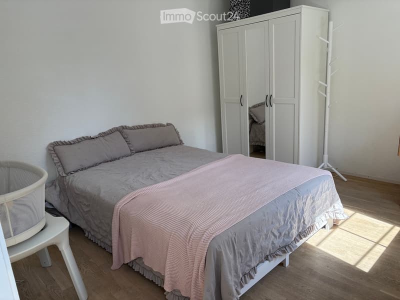 A well-lit bedroom with a double bed, grey bedding, a white wardrobe, a small white stool, and a baby chair in the corner.