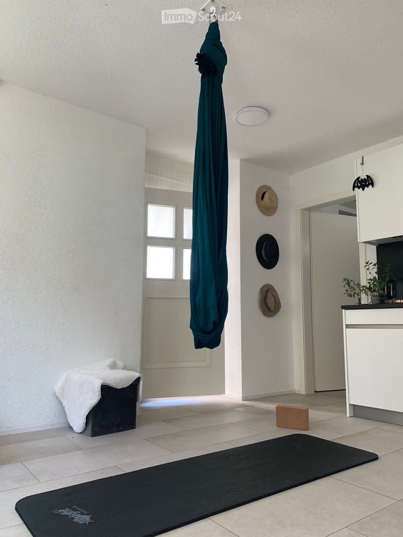 A clean, minimalist hallway with white walls, tiled floor, and several items including a hanging umbrella, hats on the wall, a small stool with a white cloth, a yoga mat, and a small counter with a plant and cup.