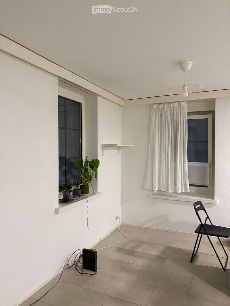 A modern, minimalist balcony with white walls, tiled floor, and large windows. Features a black folding chair, a small shelf with potted plants, a ceiling fan, and a curtain on one window.