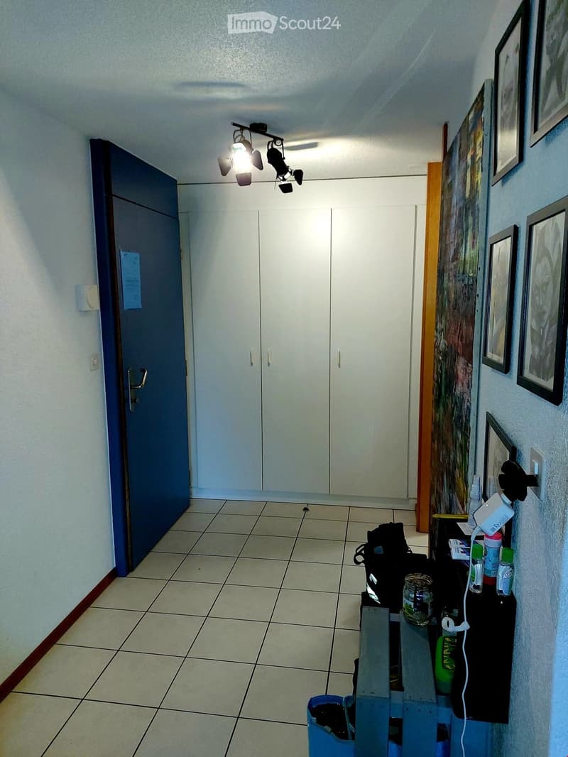 A clean, well-lit hallway with tiled flooring, white walls, and a closed door on the left. A small table with various items, including a backpack, bottles, and a jar, is positioned against the right wall. Several framed pictures and artwork are displayed