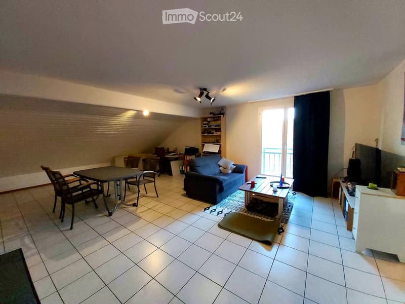A well-lit living room with a blue sofa, coffee table, dining table with chairs, and a shelf with books and decorative items. The room features tiled flooring and large windows with blinds.