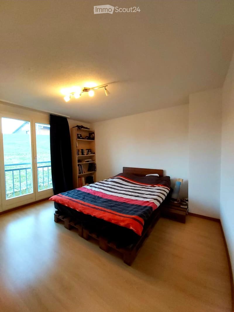 Bedroom with a wooden bed, nightstand, shelf, and balcony access. Light-colored walls and carpeted floor.