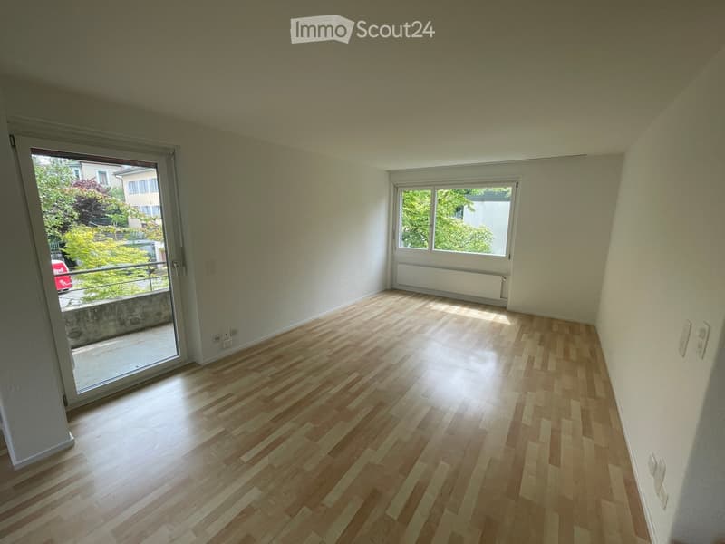 Empty room with wooden flooring, white walls, large windows, and a sliding glass door leading to a balcony.