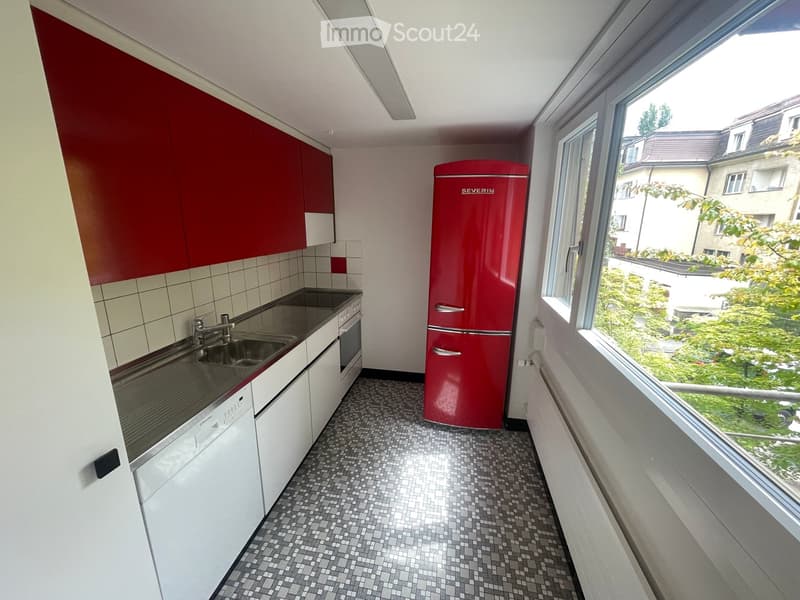 Modern kitchen with white and red cabinets, a stainless steel sink, a red refrigerator, and a dishwasher. The room features a tiled floor and large windows offering a view of the outside.