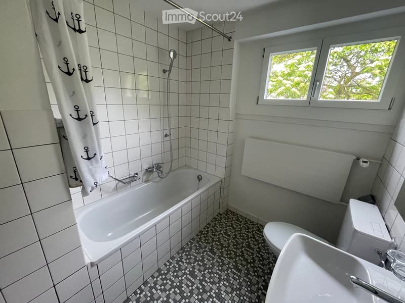 Bathroom with white tiled walls, a bathtub, toilet, sink, and a shower curtain with anchor designs. The room has a window with a view of greenery outside.