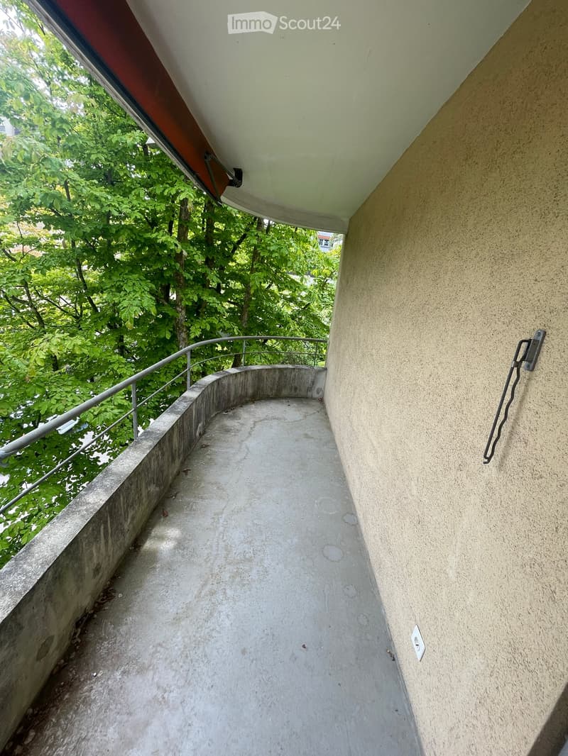 A narrow, curved balcony with a metal railing, overlooking a green, leafy area. The balcony has a concrete floor and beige walls.