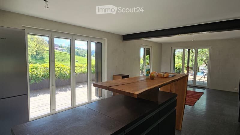 Modern kitchen with large sliding glass doors, wooden table, refrigerator, and a view of a green landscape.