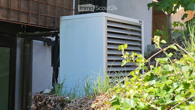 A white air conditioning unit is mounted on a wall, surrounded by greenery and plants. The unit has horizontal slats on its side and is positioned near a building with a metal railing above it.