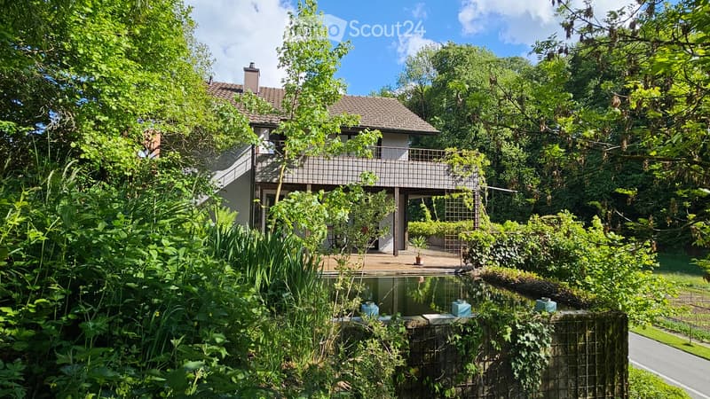 A two-story house with a tiled roof, surrounded by lush greenery and a small pond in the foreground. The house features a balcony and is partially obscured by trees and plants.