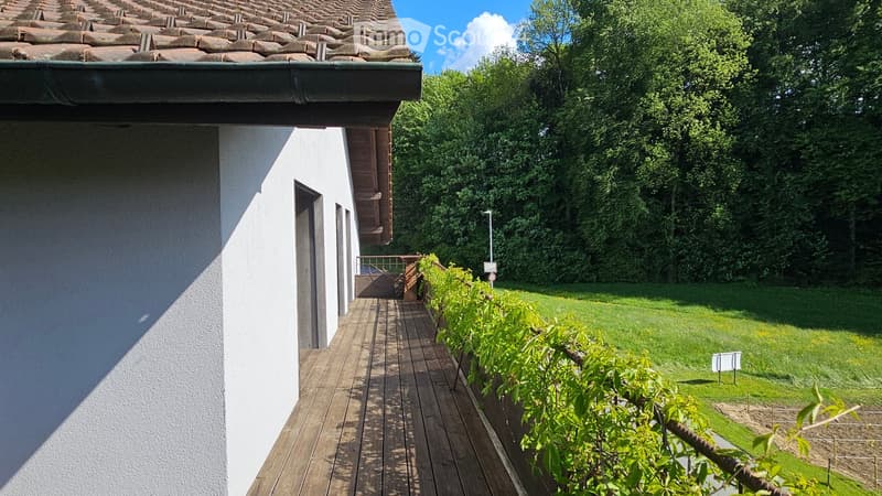 A wooden deck extends from a white house with a tiled roof, surrounded by a well-maintained lawn and lush greenery.