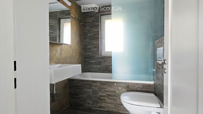 Modern bathroom with dark tiled walls, white sink, bathtub, and toilet. Features a frosted glass shower enclosure and a window for natural light.