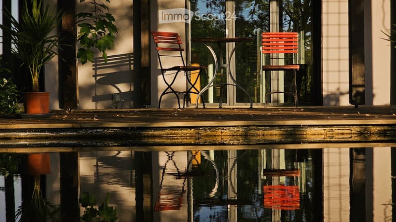 A wooden deck area with a small table and two chairs, a potted plant, and a reflective water surface below.