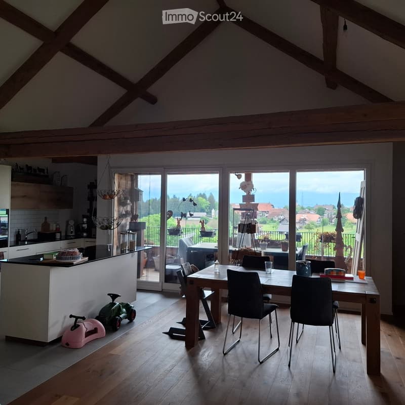 Furnished Room. Bright, open-plan room with wooden floors, a kitchen area, a dining table with chairs, and large windows offering a scenic view of the countryside.