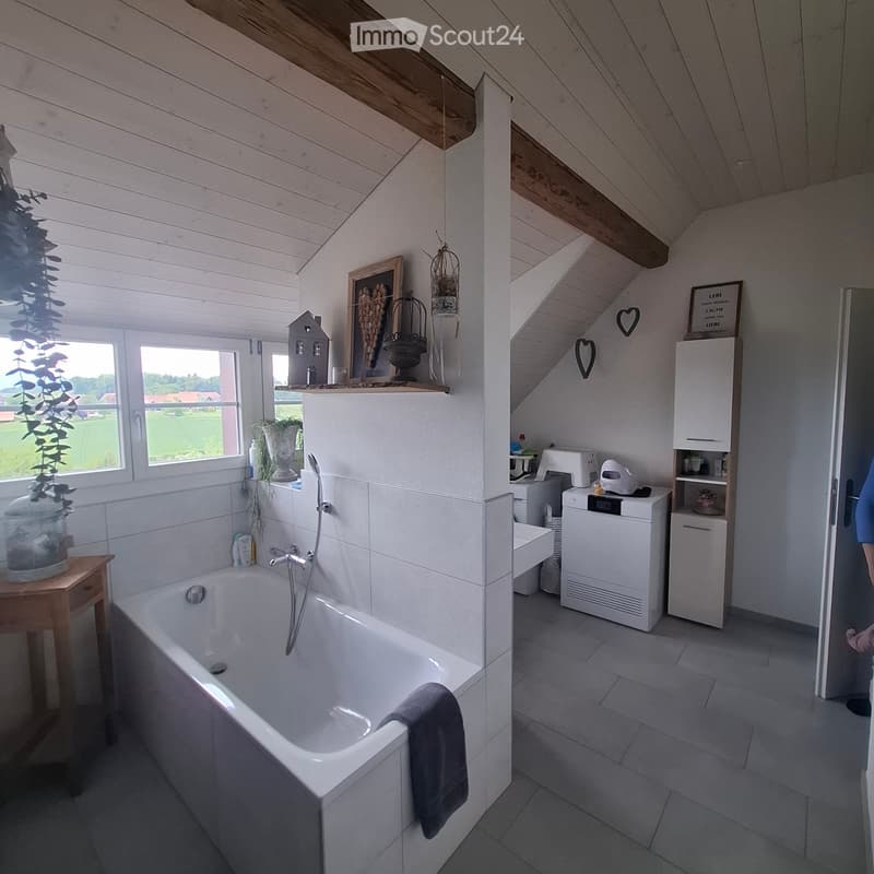 Bathroom. Bright bathroom with a white bathtub, tiled walls, wooden ceiling beams, and a washing machine. Features include a small wooden table with a vase, framed picture, and decorative items on the windowsill.