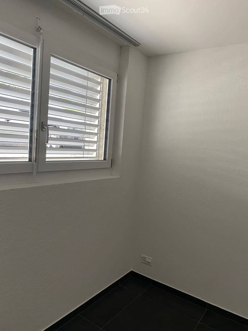 Empty room with white walls, dark tiled floor, and a window with white blinds. No furniture or equipment present.