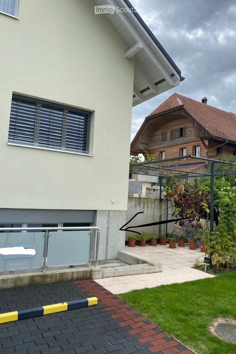 A modern two-story house with a raised ground floor, featuring a concrete staircase leading to a patio area with potted plants and a metal railing. The house has white walls, large windows with blinds, and a red-tiled roof visible in the background.