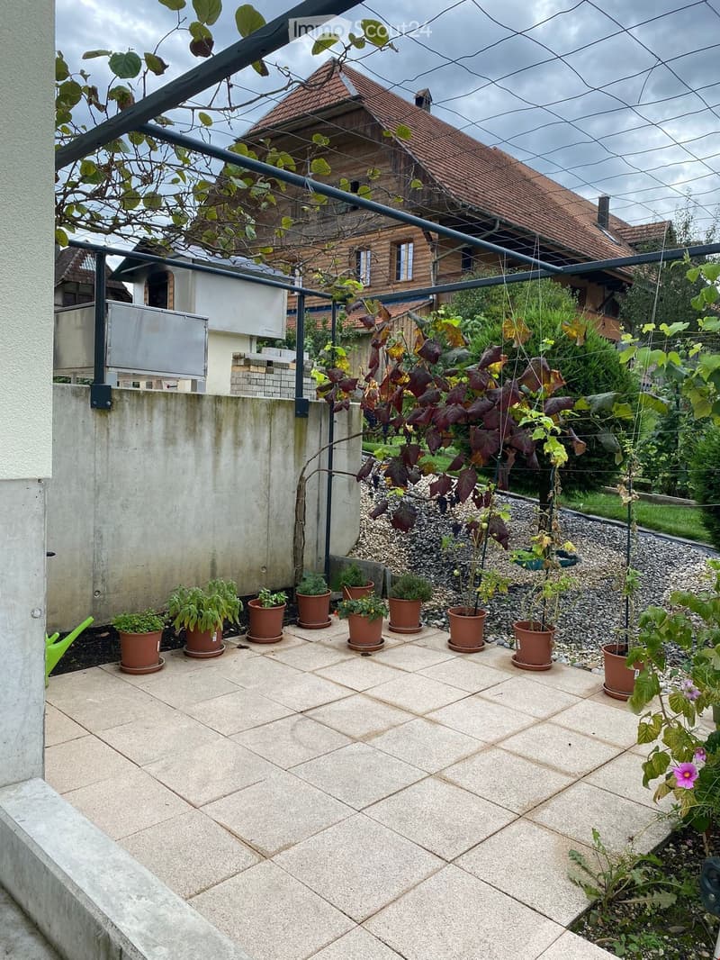 A tiled patio area with several potted plants, a metal pergola, and a view of a wooden house with a tiled roof in the background.