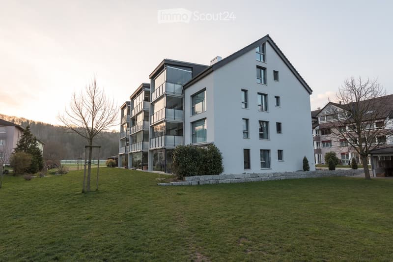 Modern multi-story residential building with white exterior, large windows, and balconies, surrounded by a well-maintained lawn and trees.