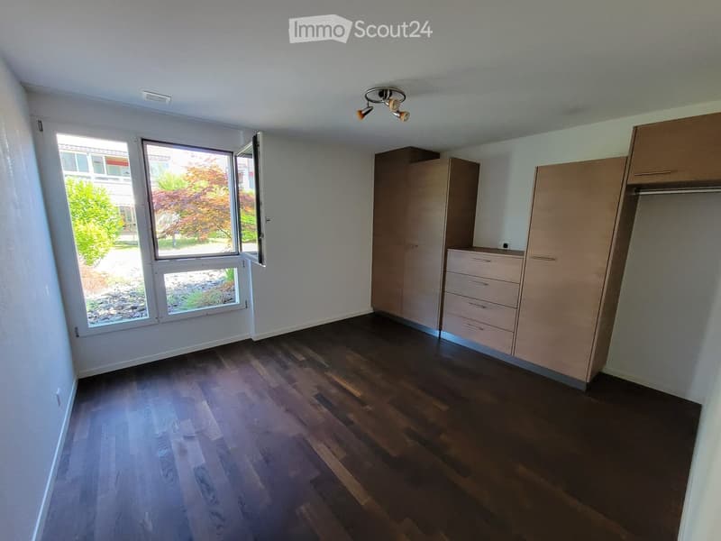 Empty room with wooden flooring, white walls, built-in closet, and large window with view of greenery.