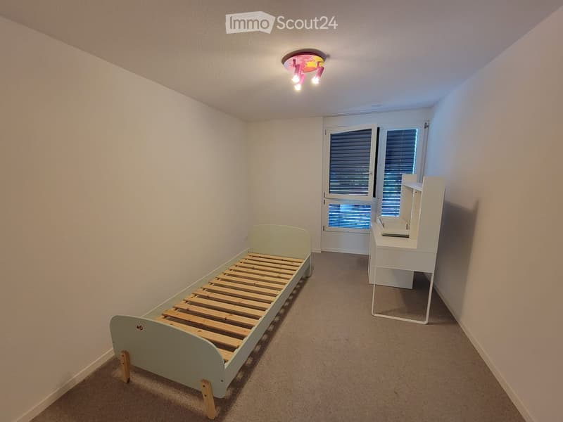 A small, unfurnished bedroom with a single bed and a nightstand, featuring a window with blinds and a door leading outside.