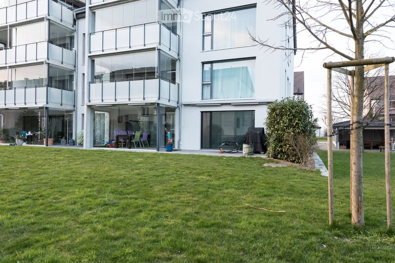 Lush green lawn in front of a modern white building with large glass windows and balconies. The building features contemporary design with sleek lines and large windows, and there is a small tree and a wooden structure in the garden area.