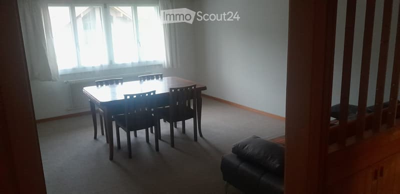 Dining area with a wooden dining table and chairs, a black leather sofa, and large windows with curtains.