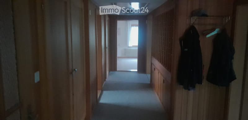 A long, narrow hallway with wooden walls and doors on both sides, leading to a room with a window at the end. Two coats are hanging on hooks on the right wall.