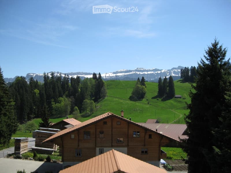A wooden house with a red-tiled roof, surrounded by green fields and trees, with snow-capped mountains in the background.