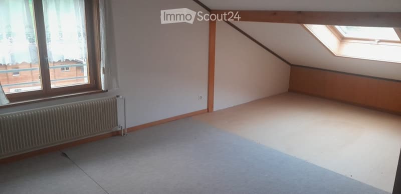 Empty attic room with sloped ceilings, large window with white curtains, radiator, and skylight.