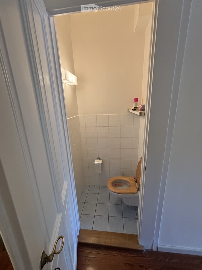 Small bathroom with white tiled walls and floor, a toilet with a wooden seat, a toilet paper holder, and a small shelf with toiletries.