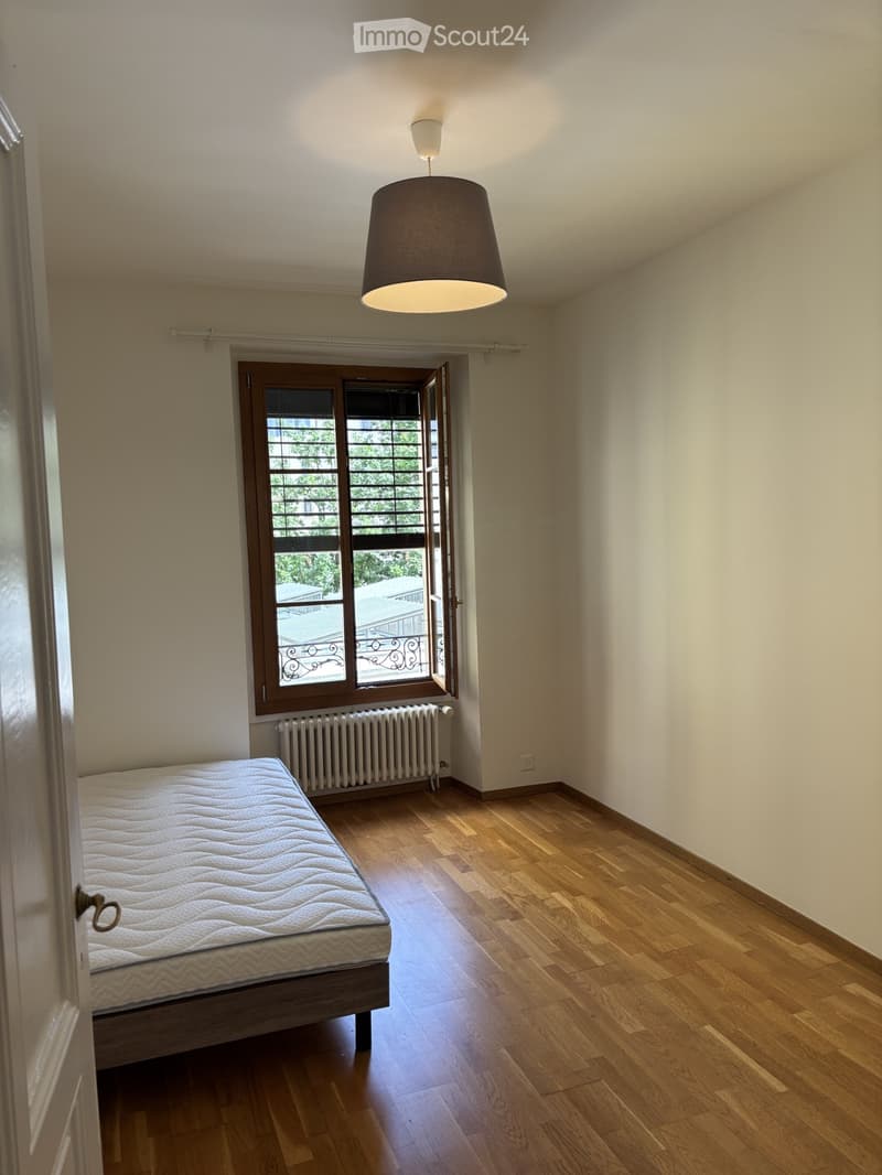 Empty bedroom with wooden flooring, a single bed, a radiator, and a hanging lamp. The room has large windows with wooden frames and a view of the outside.