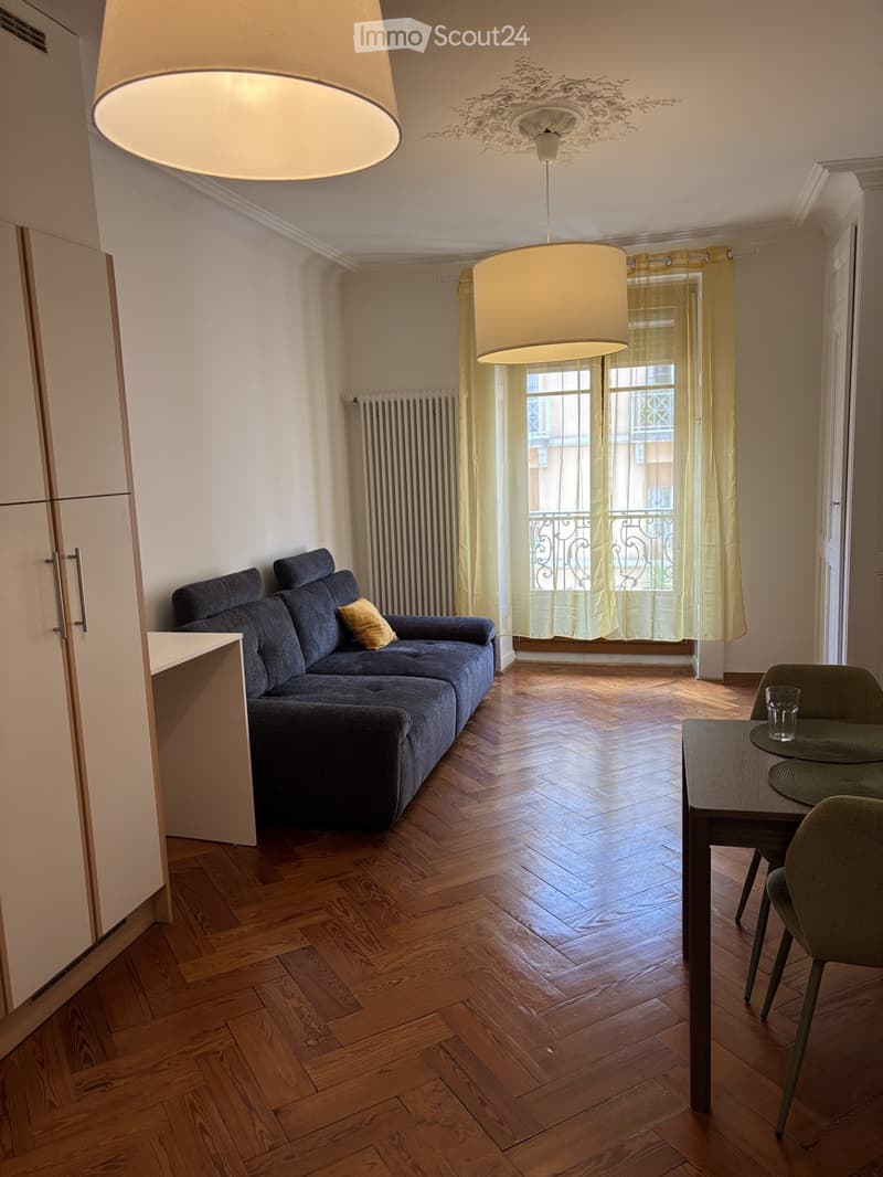 Living room with wooden flooring, a dark blue sofa, a small table with chairs, and a cup on the table. There is a large window with yellow curtains and a radiator below it.