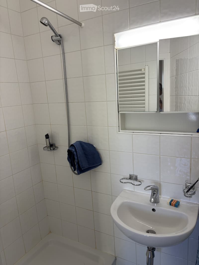 Clean bathroom with white tiled walls and floor, a white sink with a silver faucet, a mirror above the sink, a towel hanging on a rack, and a small soap bar on the sink.