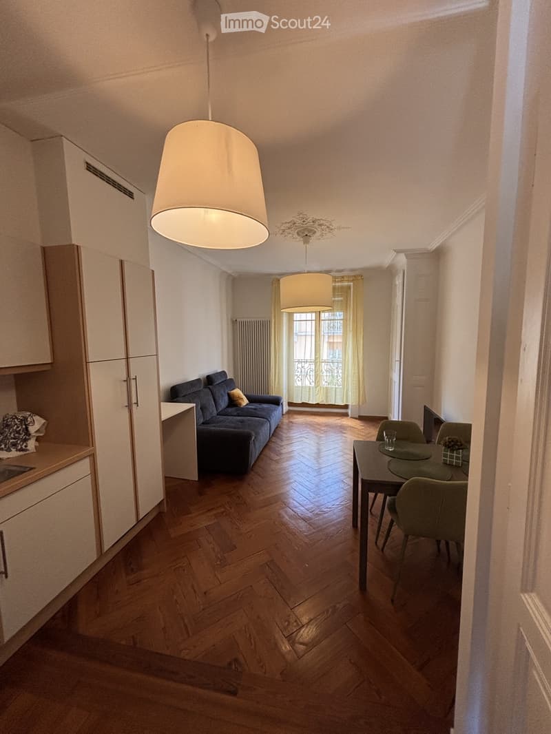 Open-plan living and dining area with wooden herringbone flooring, a blue sofa, a dining table with chairs, and a kitchen area with cabinets and a lamp.