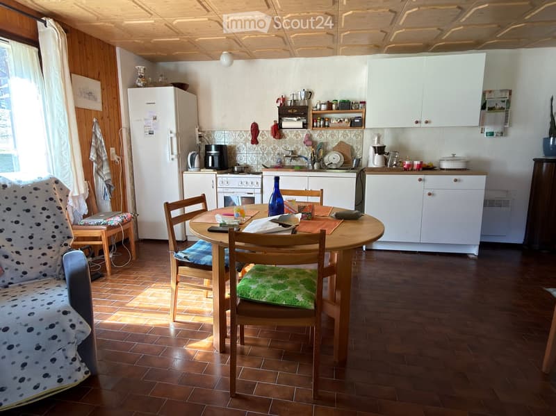A cozy kitchen and dining area with wooden flooring, white cabinets, a round wooden dining table with four chairs, a sofa with a patterned cover, and various kitchen appliances including a refrigerator, oven, and coffee maker.